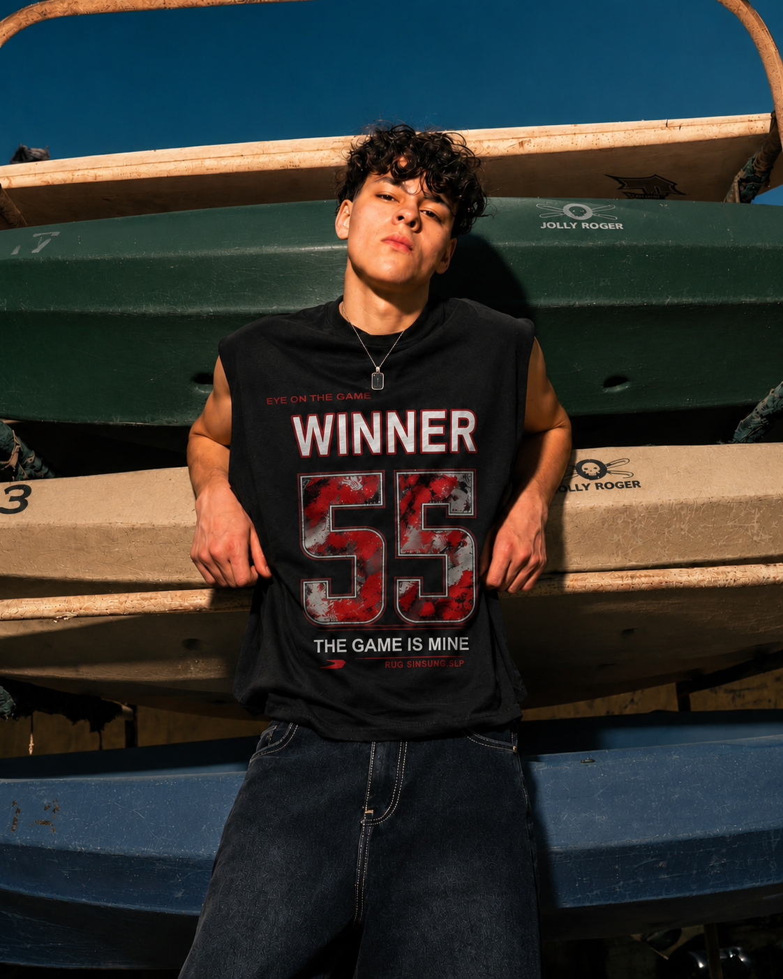 A young man wearing a black sleeveless t-shirt with a red and white graphic print that reads 'WINNER 55 THE GAME IS MINE', standing in front of stacked boats under a blue sky.