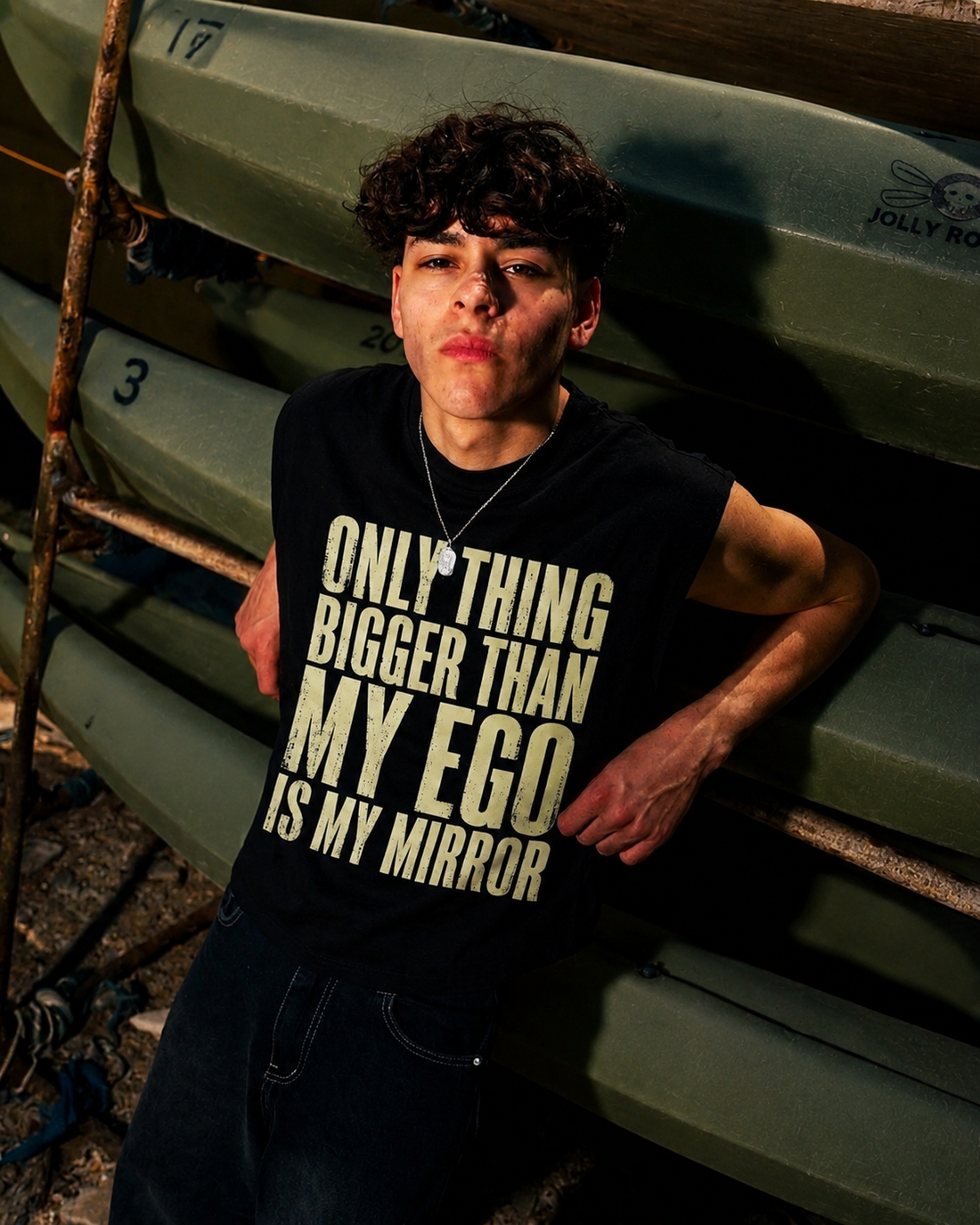 A young man sitting outdoors on a dock or boat area, wearing a sleeveless black shirt with large cream text that reads 'ONLY THING BIGGER THAN MY EGO IS MY MIRROR'.