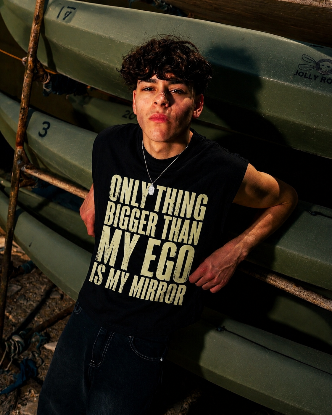 A person wearing a black sleeveless top with bold cream text reading 'ONLY THING BIGGER THAN MY EGO IS MY MIRROR', posed in front of stacked green boats.