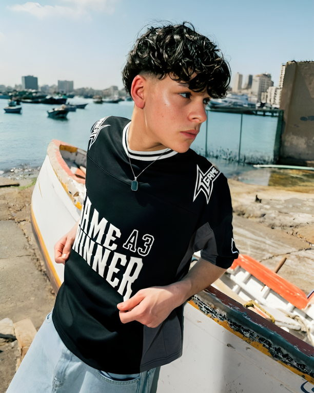 Person wearing a black t-shirt with text, standing by a waterfront with boats and buildings in the background.