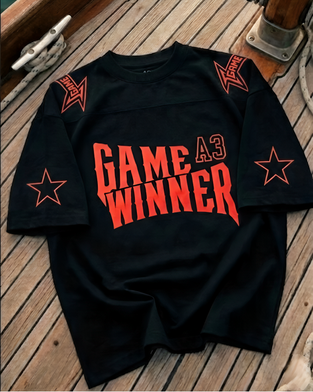 Black t-shirt with 'GAME A3 WINNER' text and red stars on a wooden deck background