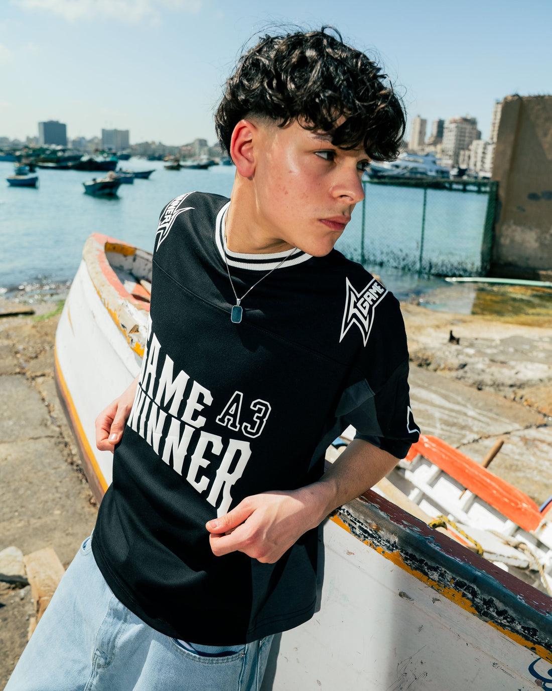 Person wearing a black 'Game Winner' t-shirt by a waterfront with boats and buildings in the background.