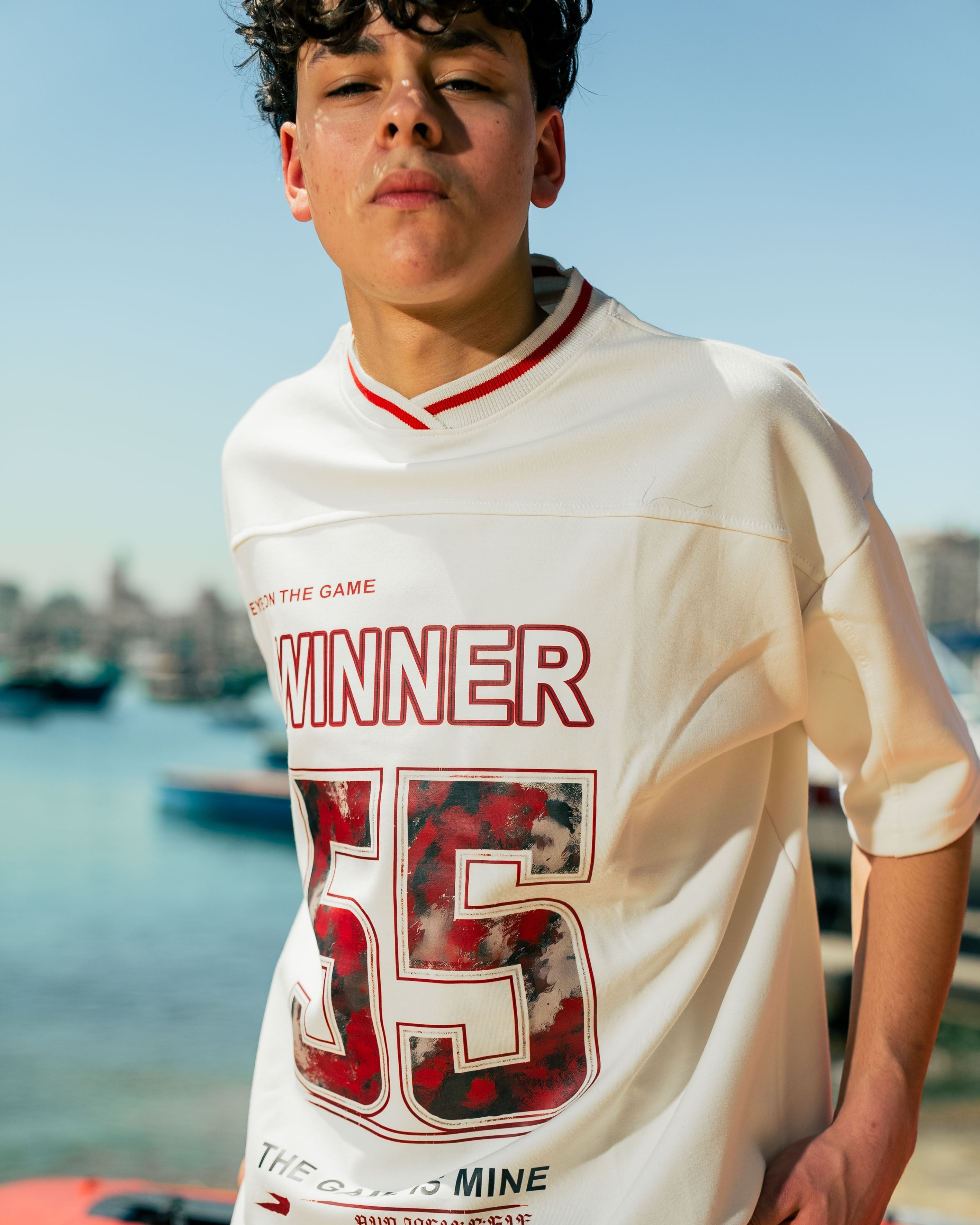 Person wearing a white t-shirt with red and black graphics and text, standing by a waterfront.