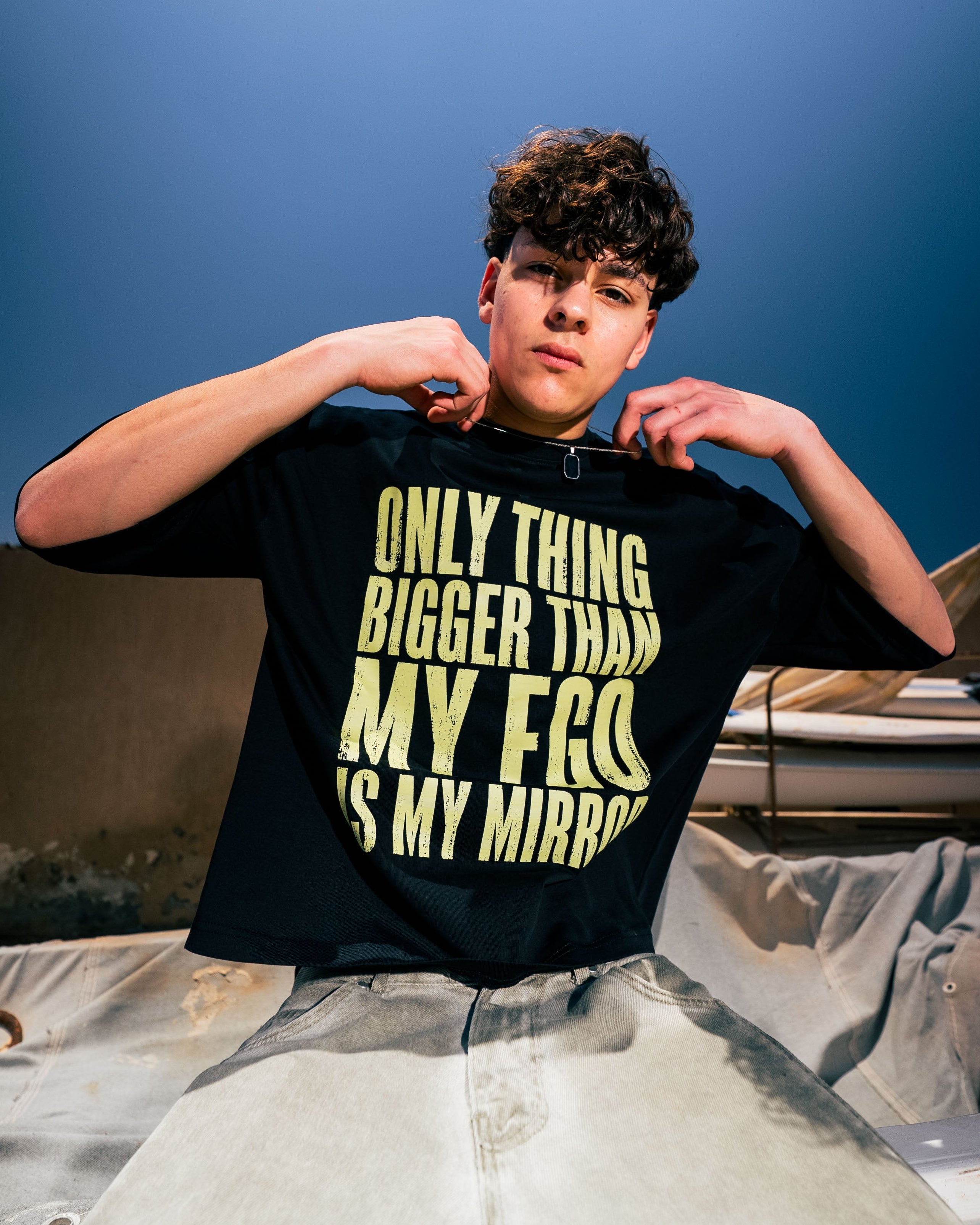 Person wearing a black t-shirt with yellow text, sitting on a rooftop with a blue sky background.