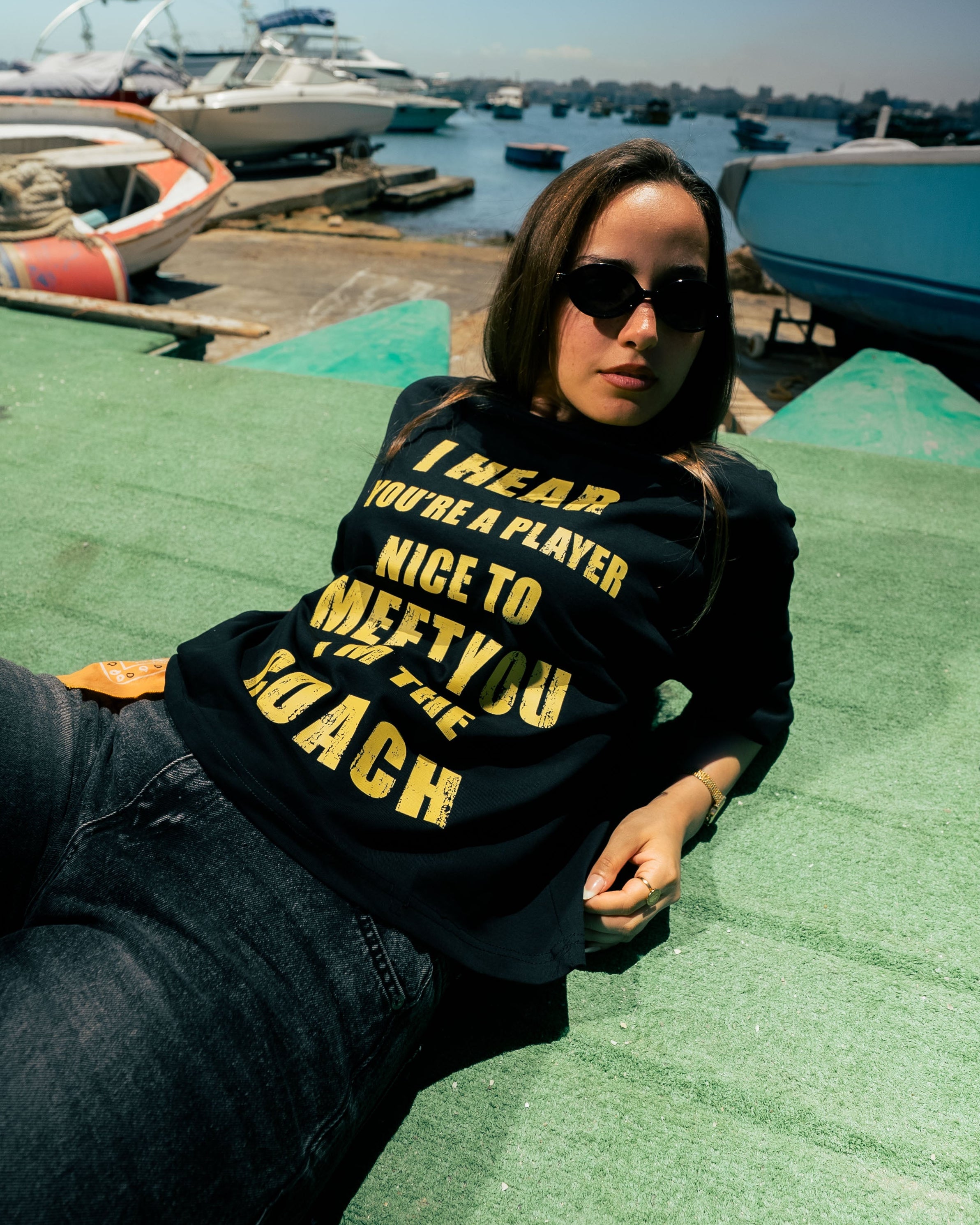 Person wearing a black sweatshirt with yellow text at a dock with boats in the background