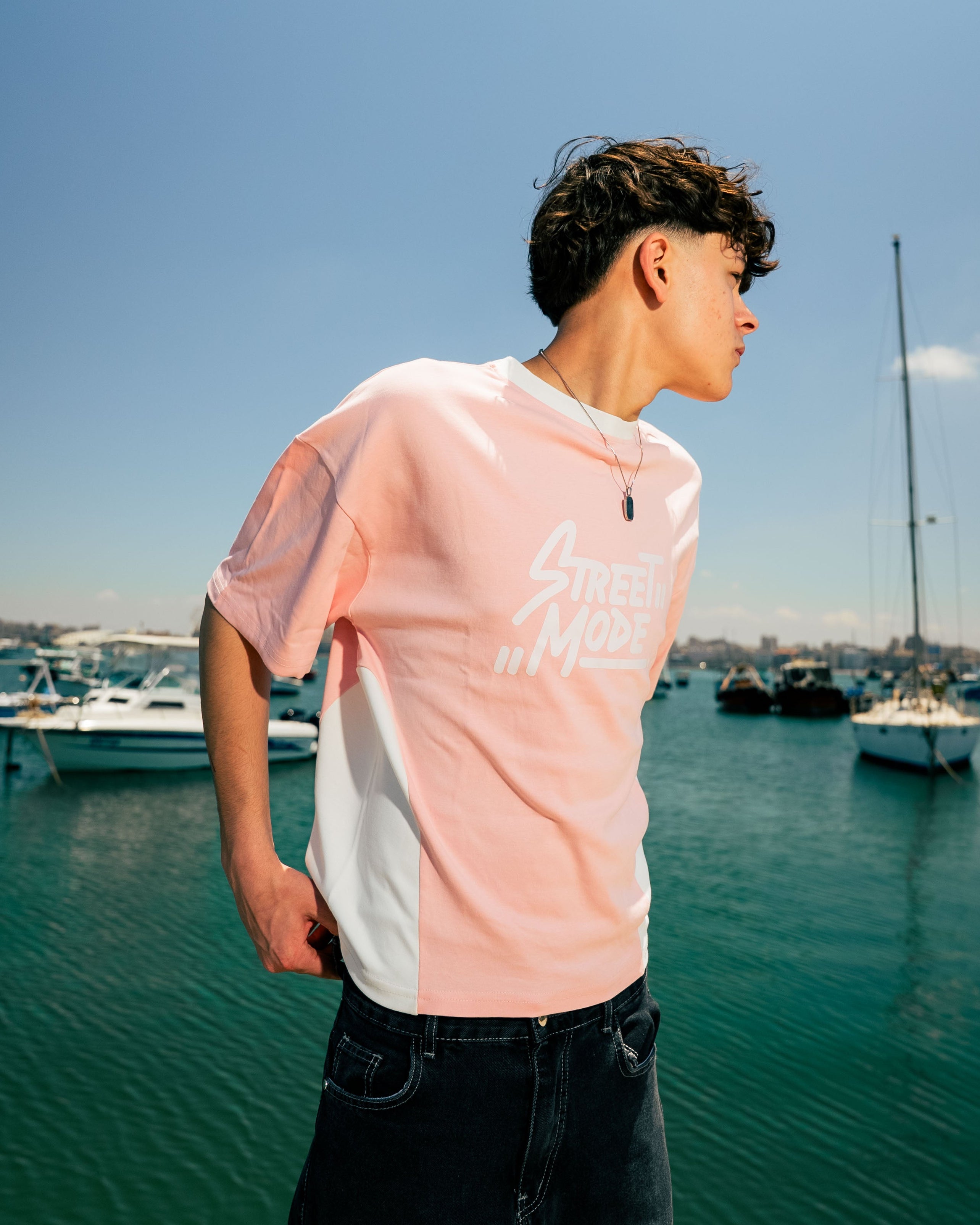 Person wearing a pink and white t-shirt by a waterfront with boats and clear sky.