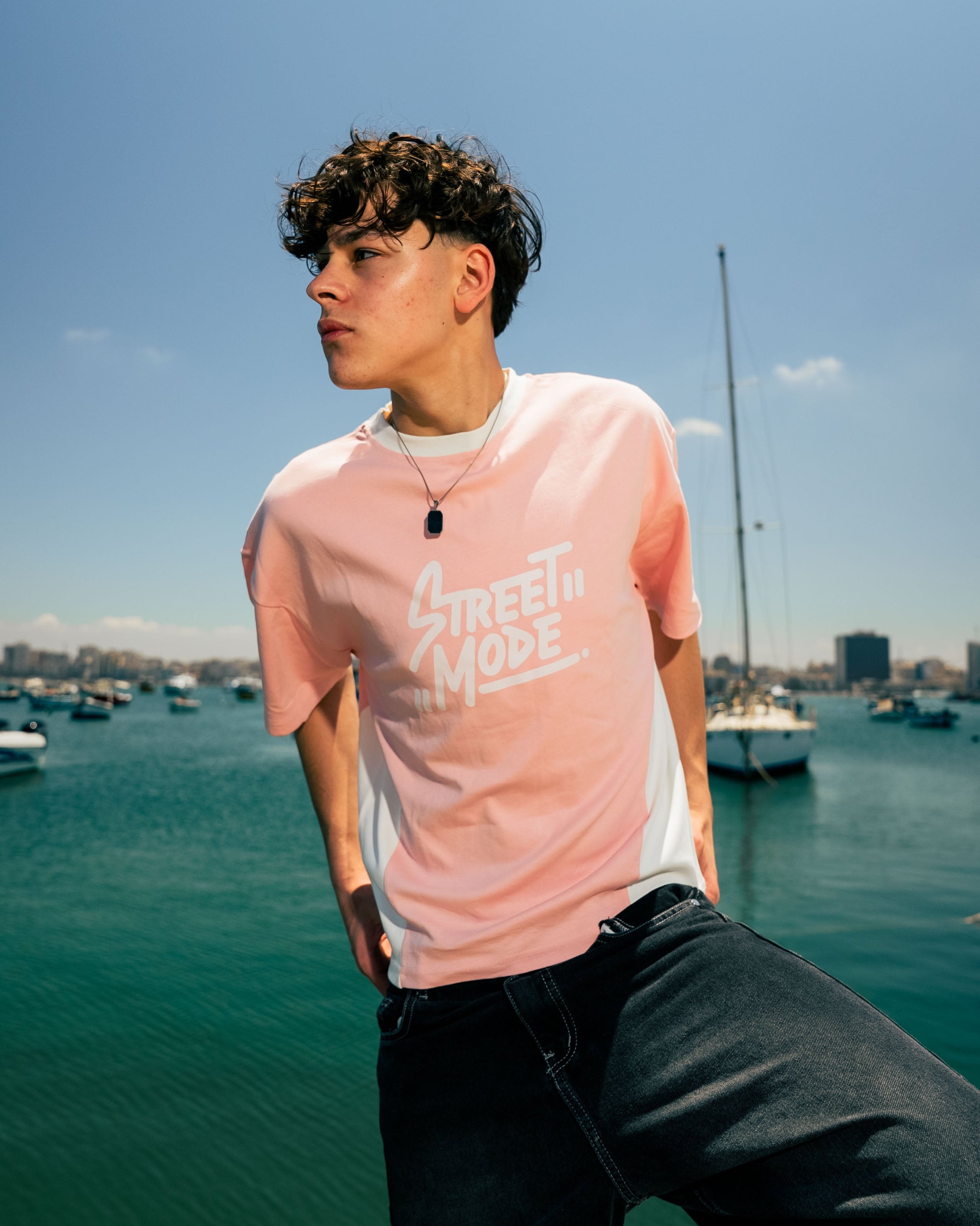 Person wearing a pink t-shirt with text, standing by a waterfront with boats and buildings in the background.