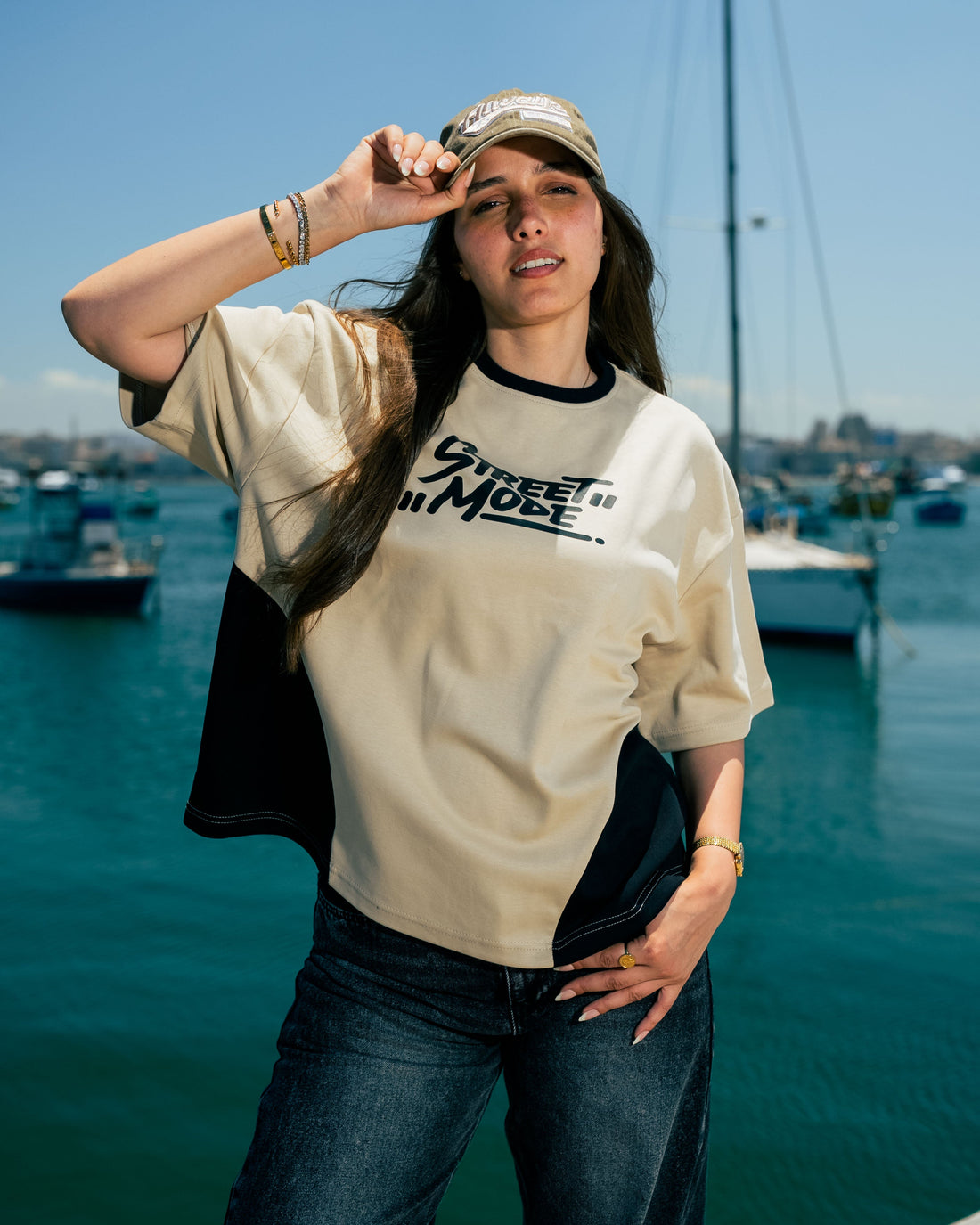 Woman wearing a beige and black shirt with text, standing by a waterfront with boats and clear blue sky.