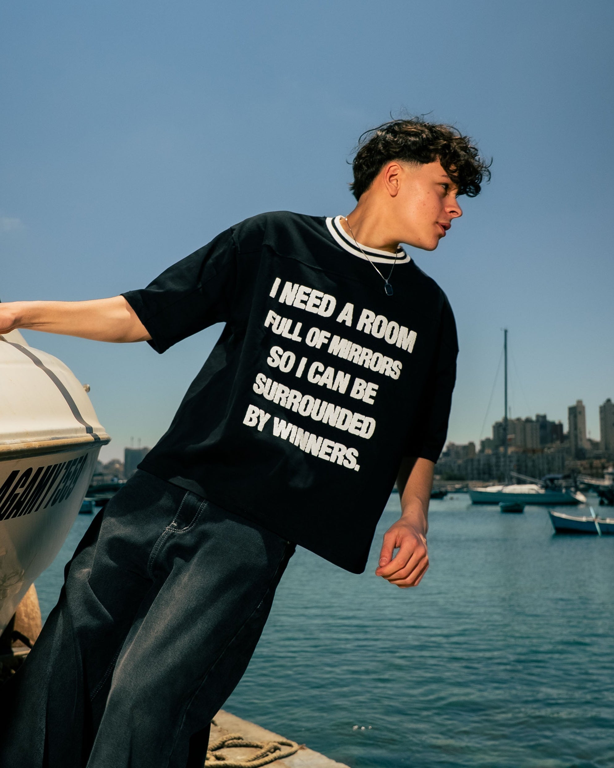 Person wearing a black t-shirt with text by a boat on a dock with water and city skyline in the background