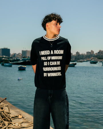 Person wearing a black t-shirt with text by a waterfront with boats and buildings in the background.