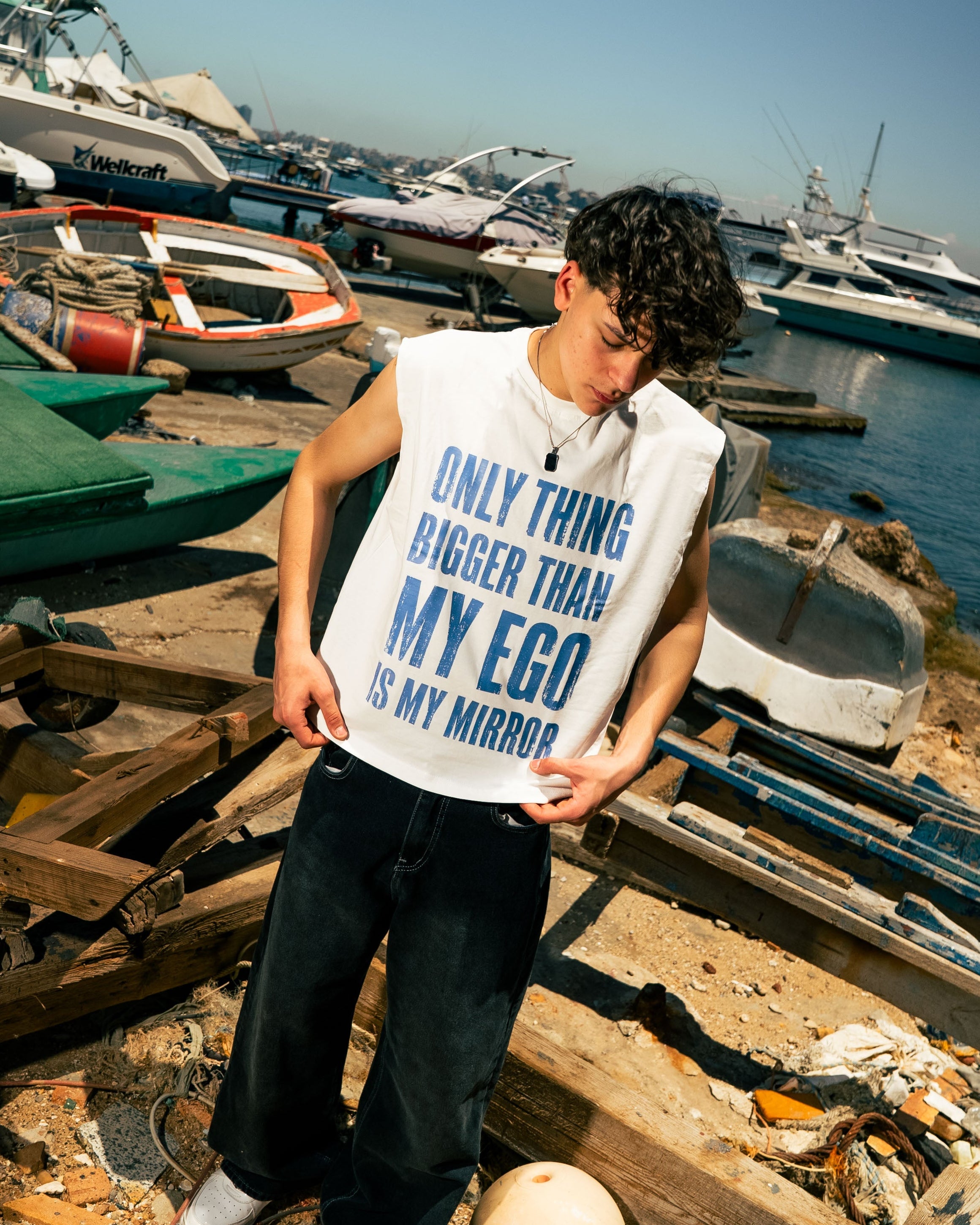 Person wearing a tank top with text by a marina with boats and wooden planks.