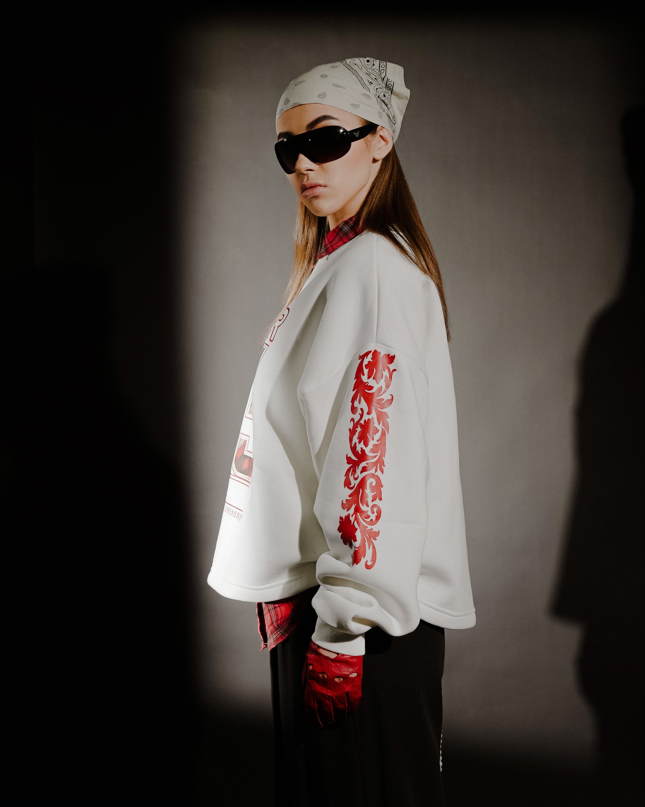 Person wearing a white sweatshirt with red designs, black pants, and sunglasses against a dark background