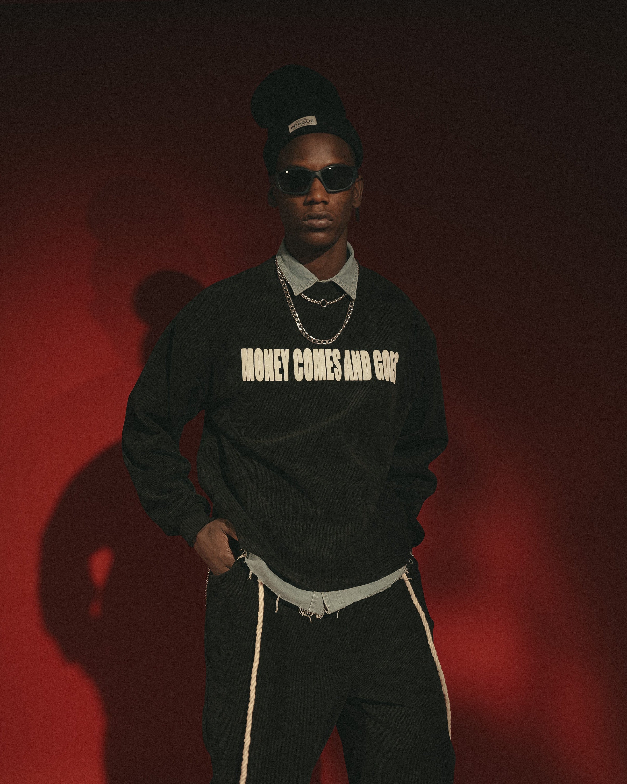 Person wearing a black sweatshirt with text, sunglasses, and dark pants against a red background