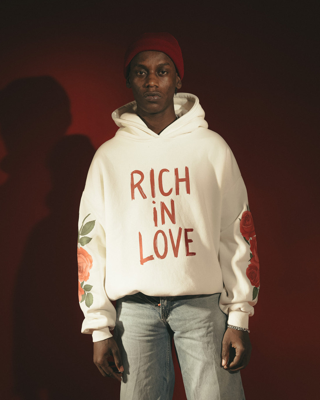 An off-white pullover hoodie with 'RICH IN LOVE' printed in a distressed red font on the chest, featuring red rose and green leaf illustrations on both sleeves, set against a solid black background.
