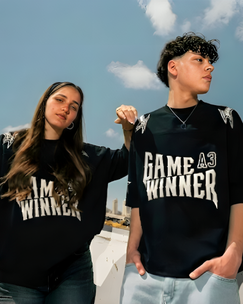 Two people wearing black t-shirts with 'GAME WINNER' text, standing outdoors with a cityscape in the background.