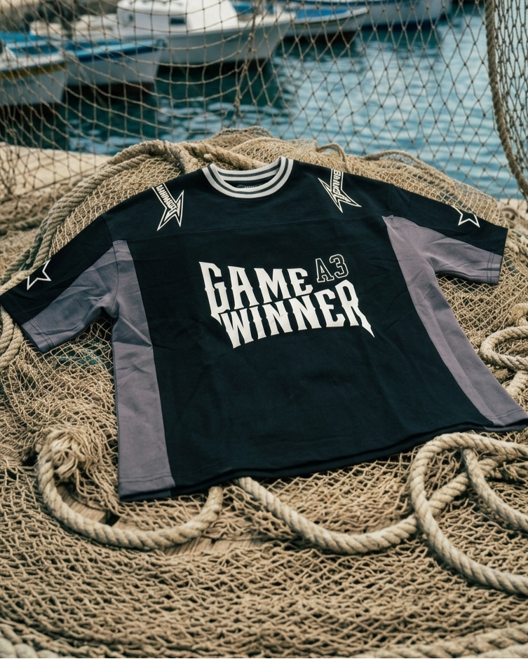 Black shirt with 'GAME WINNER A3' text on a textured surface with boats in the background