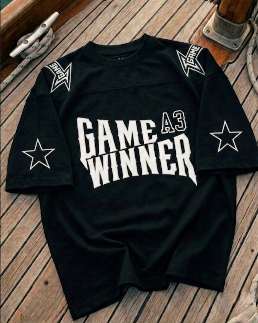 Black t-shirt with 'GAME A3 WINNER' text and star designs on a wooden surface.