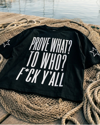 Black t-shirt with white text on a wooden surface with ropes and water in the background