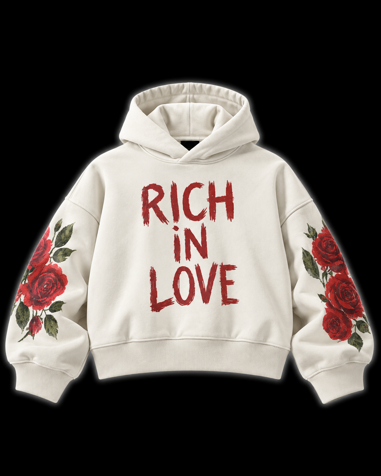 An off-white pullover hoodie with 'RICH IN LOVE' printed in a distressed red font on the chest, featuring red rose and green leaf illustrations on both sleeves, set against a solid black background.