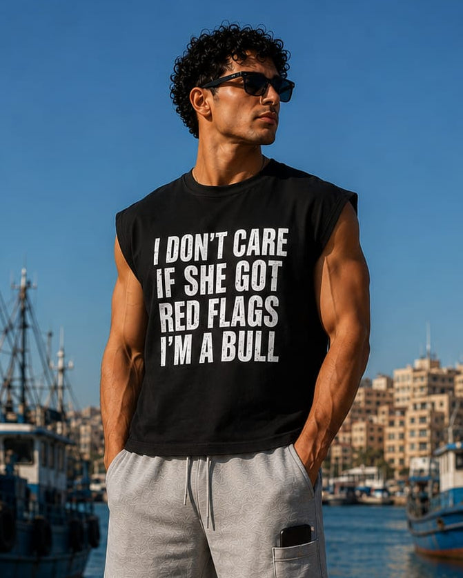 A man standing on a dock in front of a harbor with boats, wearing a black sleeveless t-shirt with bold white text that reads 'I DON'T CARE IF SHE GOT RED FLAGS I'M A BULL', paired with light gray shorts, white socks, and white sneakers.