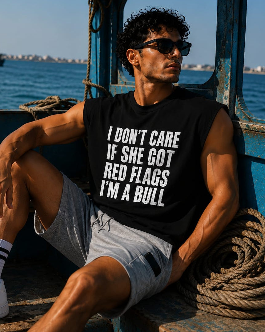 A man sitting on the wooden steps of a blue boat wearing a black sleeveless t-shirt with bold white text that reads 'I DON'T CARE IF SHE GOT RED FLAGS I'M A BULL'.