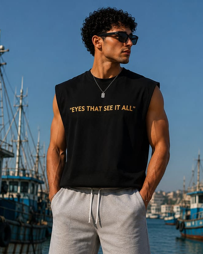 A man stands on a dock by the water wearing a black sleeveless muscle tank top with gold text that reads 'EYES THAT SEE IT ALL', light gray drawstring shorts, white socks, and white sneakers. Fishing boats and blue water are visible in the background.