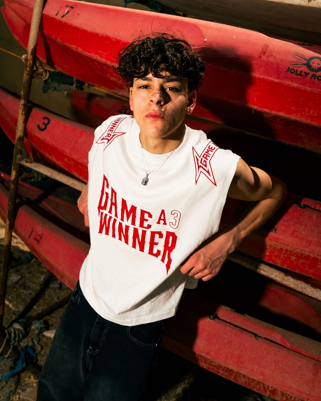 A person wearing a white sleeveless graphic T-shirt with red text reading 'GAME A WINNER' and red star graphics on the shoulders, posed in front of stacked red canoes.