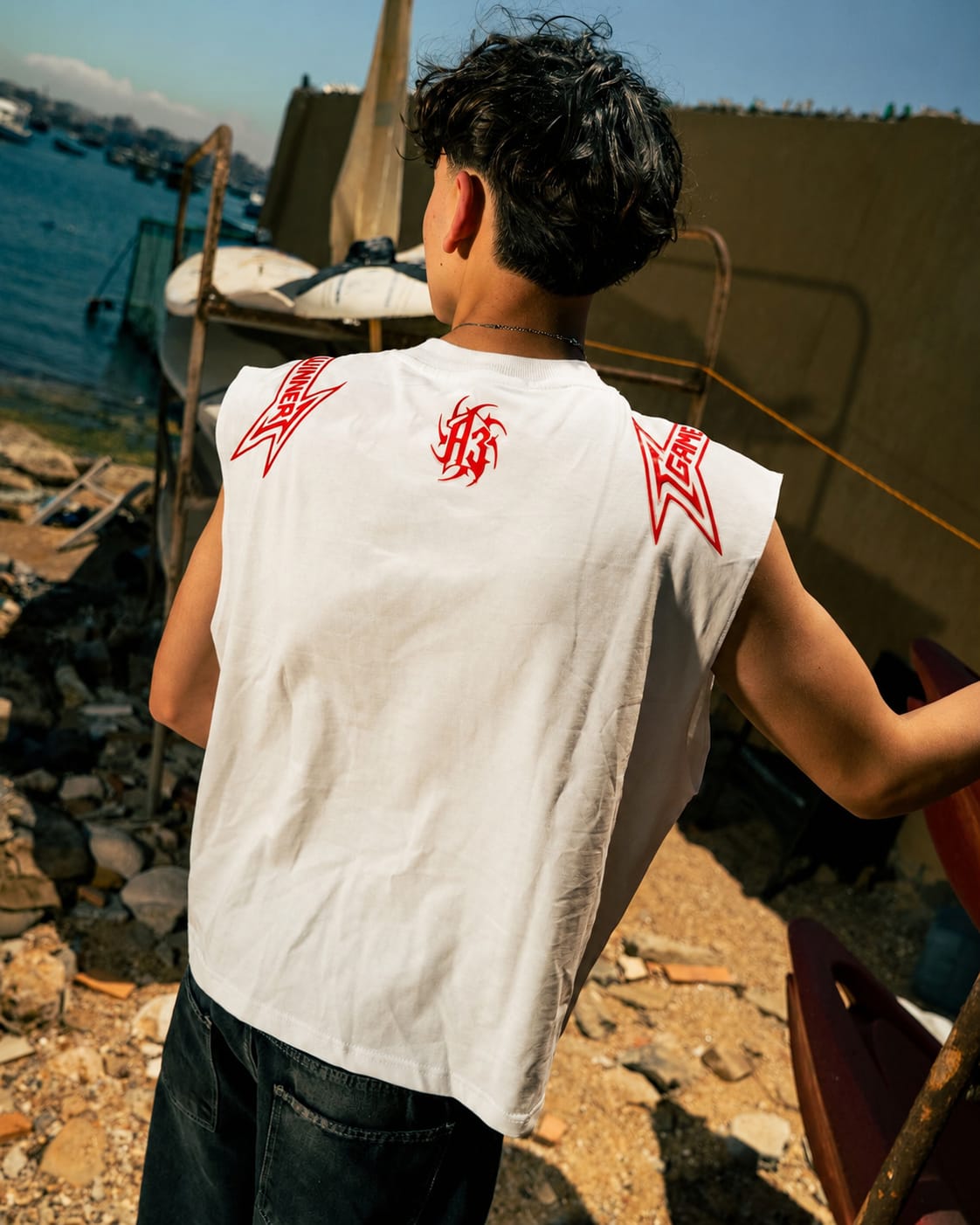 A man viewed from behind wearing a sleeveless white t-shirt with red graphic designs on the shoulders and back, set against a rustic outdoor background near water and boats.