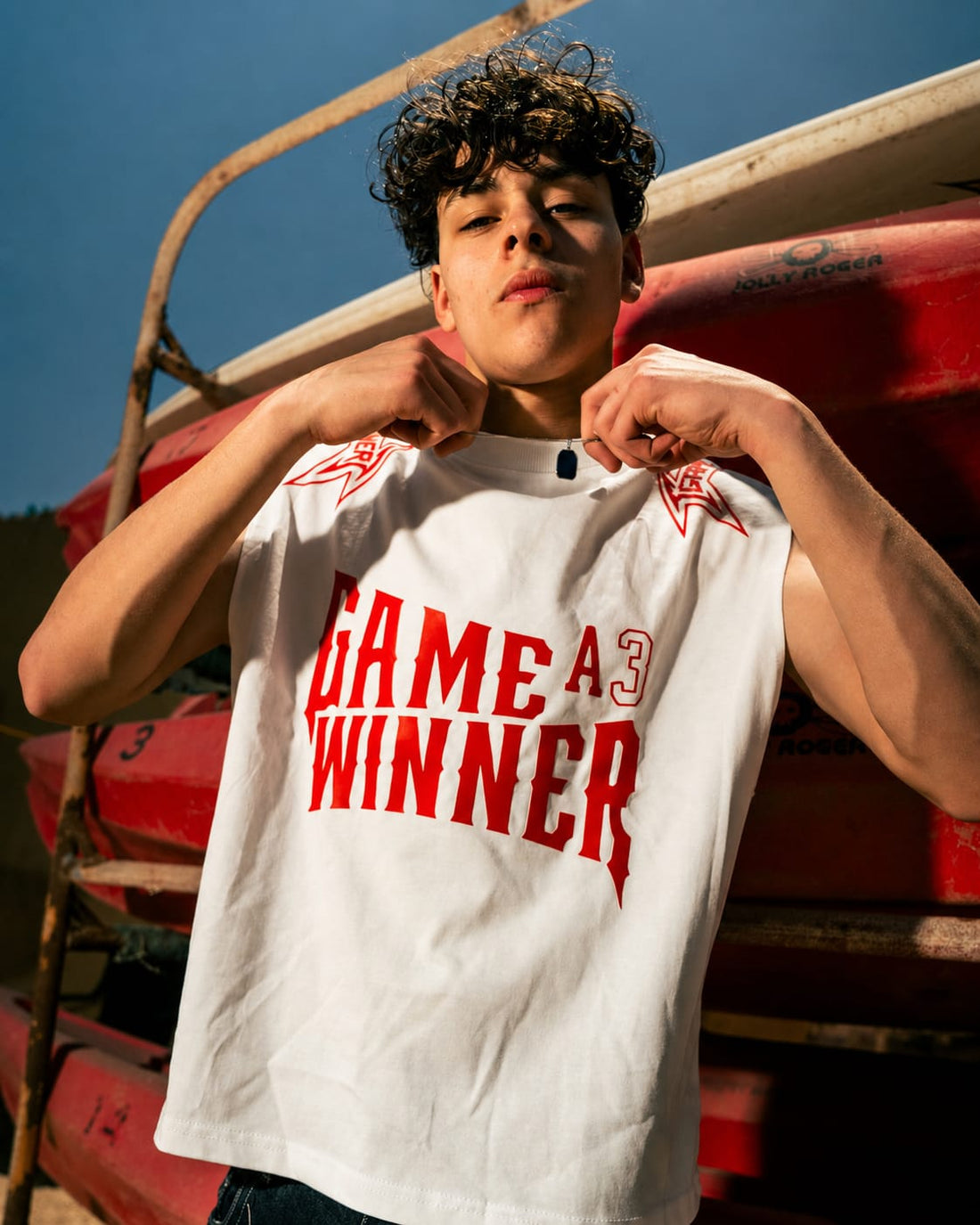 A person wearing a white sleeveless graphic T-shirt with red text reading 'GAME A WINNER' and red star graphics on the shoulders, posed in front of stacked red canoes.