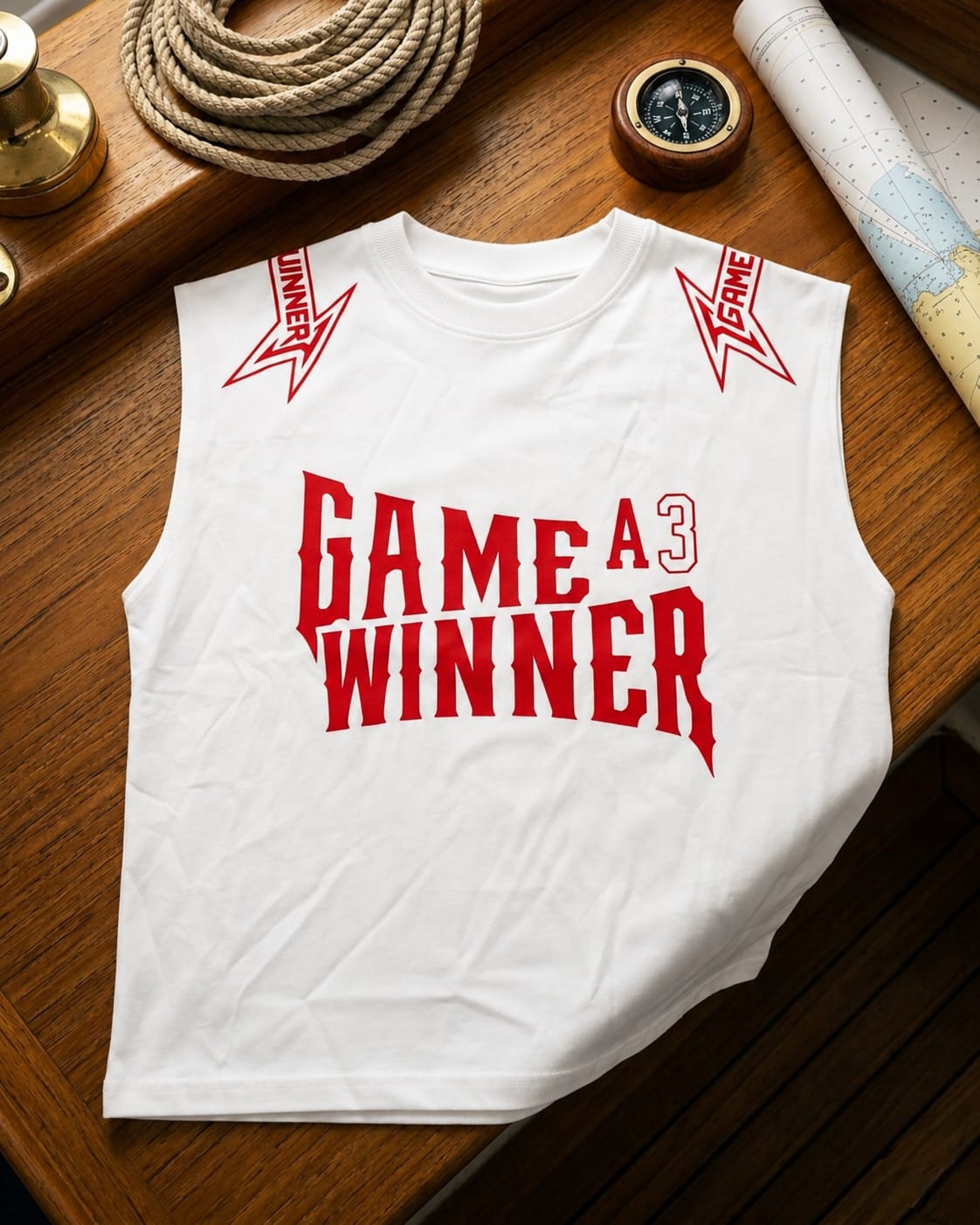 A white sleeveless crop top with red graphic text reading 'GAME A3 WINNER' and red lightning-bolt style 'WINNER' graphics on the shoulders, laid flat on a wooden surface with nautical props around it.