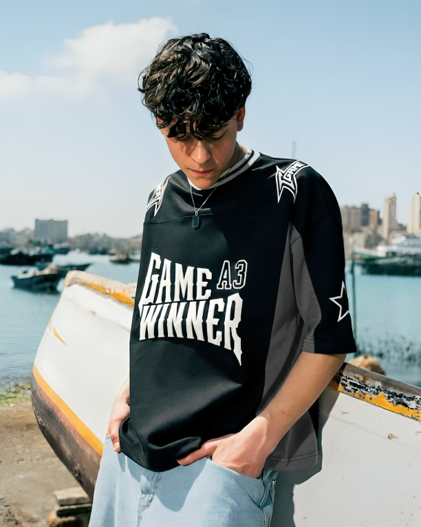 Person wearing a black sports jersey with 'Game Winner' text, standing by a waterfront with boats and buildings in the background.