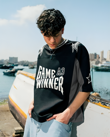 Person wearing a black sports jersey with 'Game Winner' text, standing by a waterfront with boats and buildings in the background.
