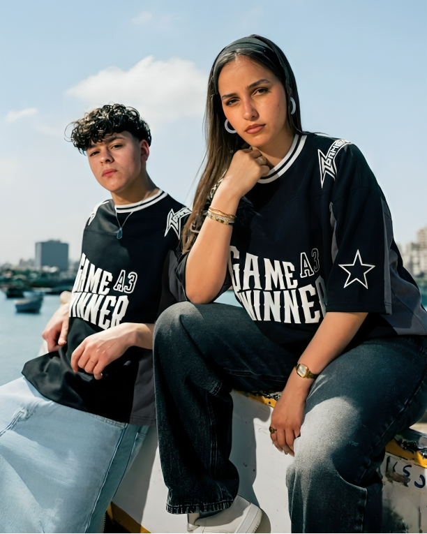 Two people wearing matching black t-shirts with text and graphics, sitting outdoors against a blue sky.