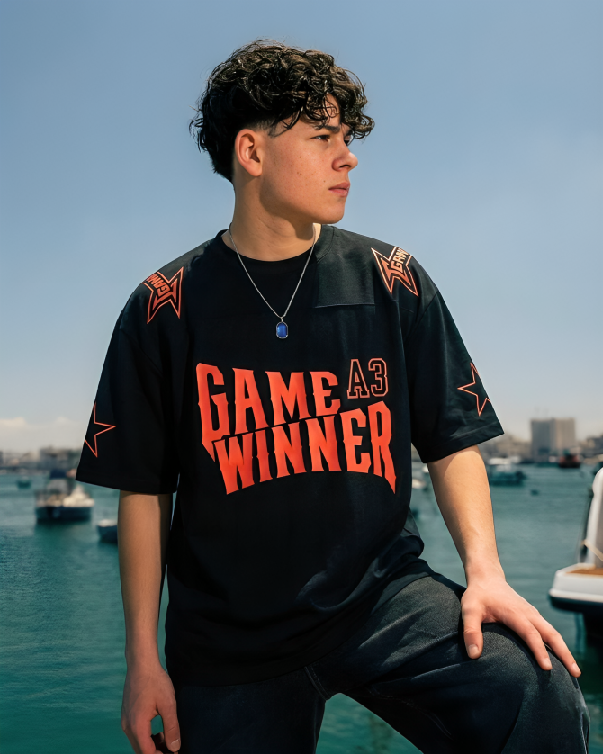 Person wearing a black t-shirt with 'GAME WINNER' text by a waterfront with boats and buildings in the background.