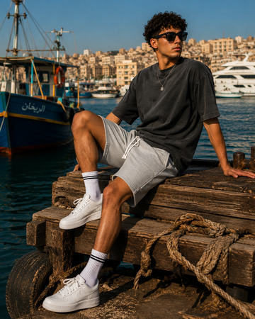 A man wearing a dark gray t-shirt, light gray shorts, white crew socks, and white sneakers sits on a wooden dock in a harbor with boats and a city skyline in the background.