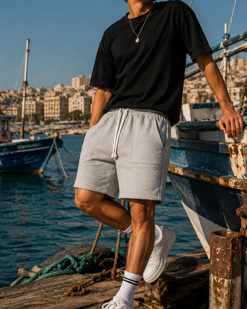 A man wearing a black t-shirt, light gray sweat shorts, white socks, and white sneakers stands on a wooden dock next to a blue boat with a harbor city in the background.